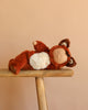A plush toy with a sleeping face, dressed in a fuzzy fox costume, lies on its back atop a wooden bench against a plain beige background—meet the snuggly Olli Ella Cozy Dinkum Doll - Fox.