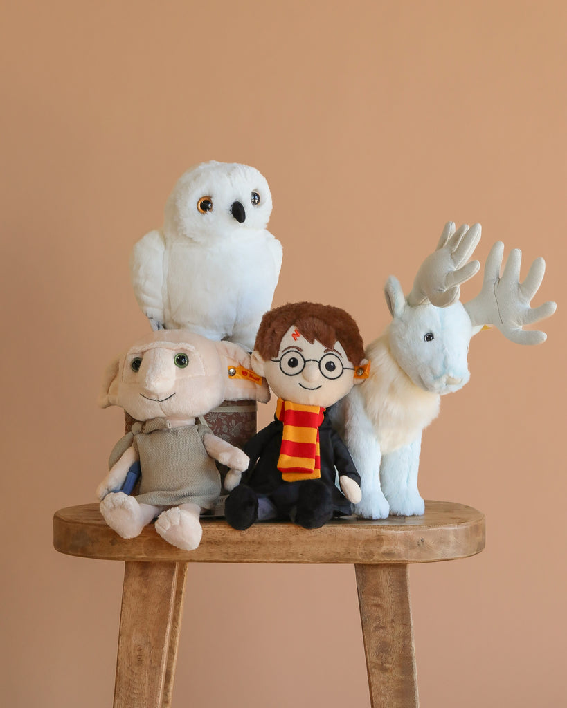 The Steiff x Harry Potter Set features four plush toys—Hedwig, Dobby, Harry Potter with glasses and scarf, and a white stag—arranged on a wooden stool against a plain beige background.