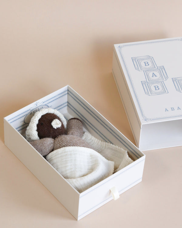 A baby gift box with a Hand-Knit Baby Doll - Cocoa and an organic cotton swaddle inside, set against a soft peach background. The box lid features a baby block design with letters.
