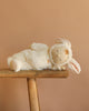 The Olli Ella Cozy Dinkum - Bunny Moppet is a collectible plush toy lying on its side in a soft white bunny costume with floppy ears, displayed on a wooden bench against a plain beige background.