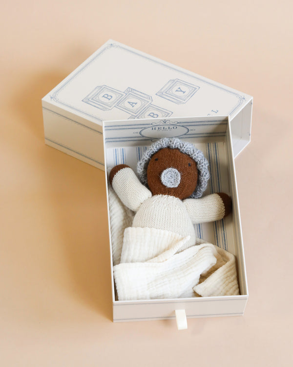 A hand-knit baby doll - caramel and an organic cotton swaddle inside an open, decorative baby gift box with "baby" and "hello" written on it, set against a soft peach background.