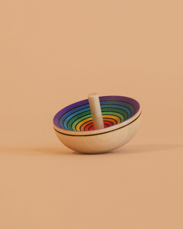 The Mader UFO Spinning Top - Purple Rainbow features vibrant concentric circles on a light peach background. Its simple wooden design and playful colors shine as it tilts, highlighting its unique charm.