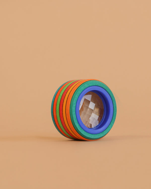 The Mader Wooden Compound Eye Lens, featuring a blue-rimmed magnifying glass and colorful maple wood rings, is displayed on a beige background.