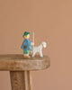 The Ostheimer The Shepherd Boy Book Set features a small handcrafted wooden shepherd boy in a green hat and blue coat with a white sheep, ideal for imaginative play, displayed on a wooden bench against a simple beige background.