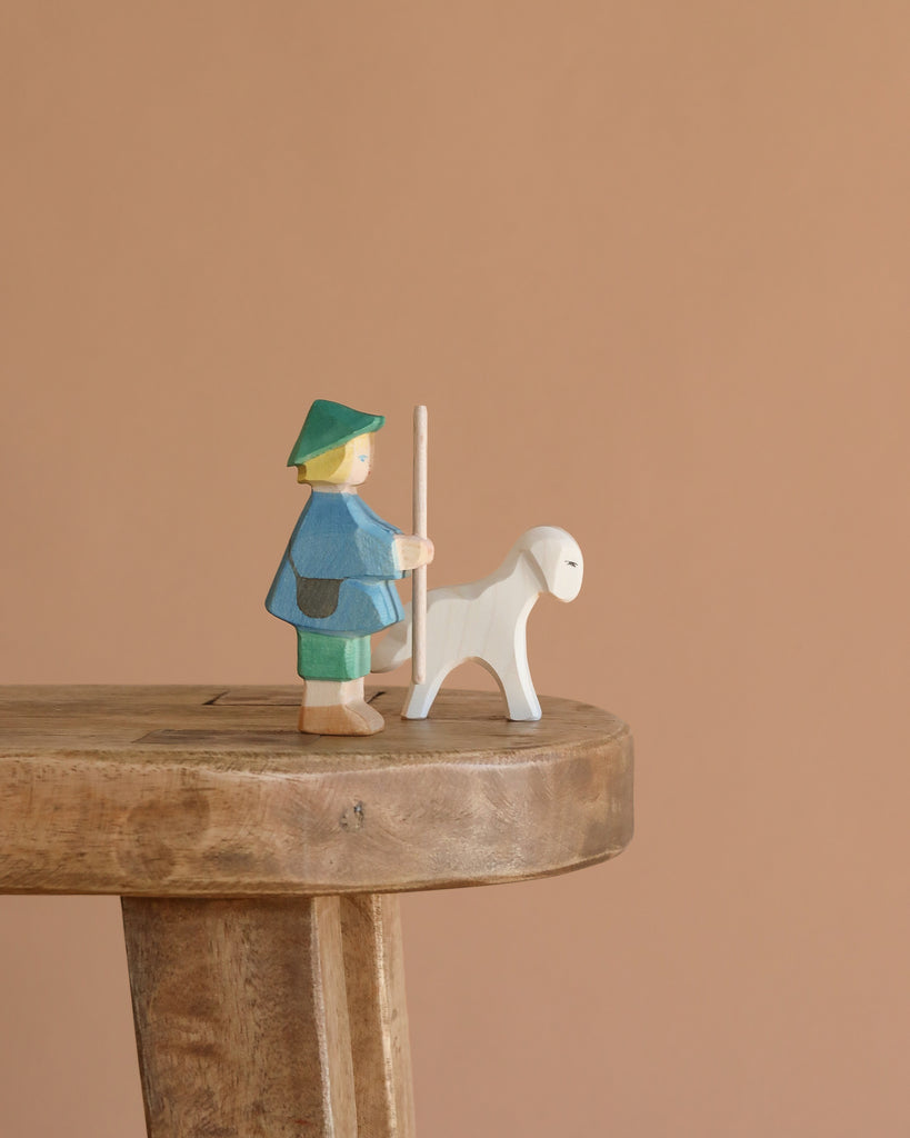 The Ostheimer The Shepherd Boy Book Set features a small handcrafted wooden shepherd boy in a green hat and blue coat with a white sheep, ideal for imaginative play, displayed on a wooden bench against a simple beige background.