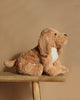 The Steiff Berno Goldendoodle Dog Plush, featuring a soft brown coat, white belly, and floppy ears, sits on a wooden stool against a beige backdrop. Its gentle expression makes it perfect for cozy cuddles.