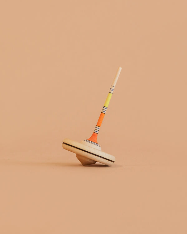 The Mader Spaghetti Spinning Top - Yellow, crafted from Maple wood with orange, yellow, and black stripes on its stem, is shown balanced upright against a plain peach-colored background.