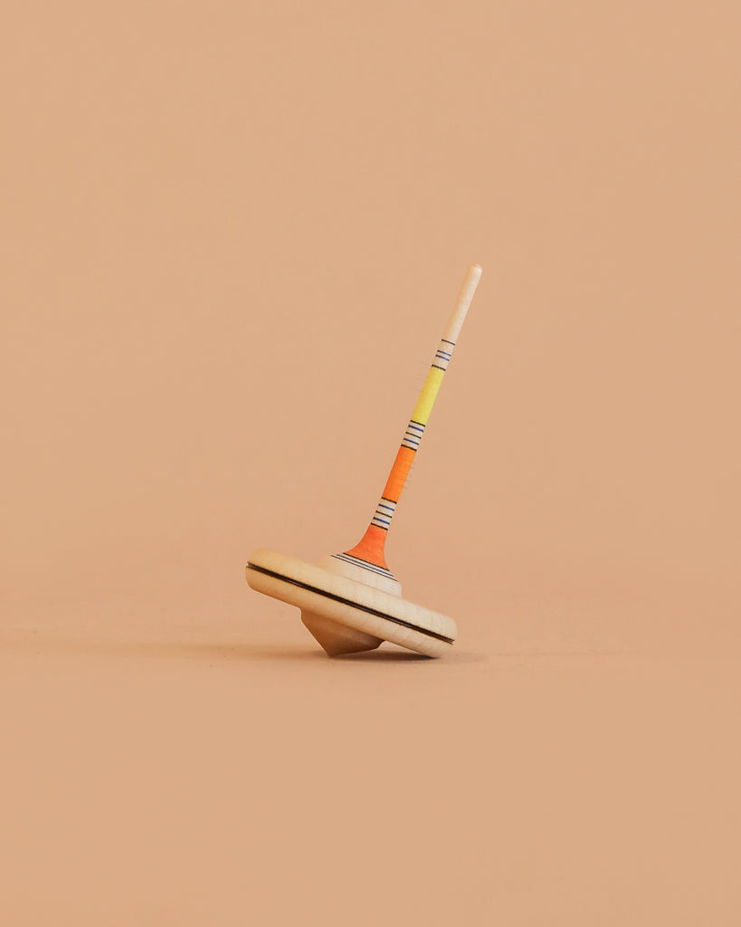 The Mader Spaghetti Spinning Top - Yellow, crafted from Maple wood with orange, yellow, and black stripes on its stem, is shown balanced upright against a plain peach-colored background.