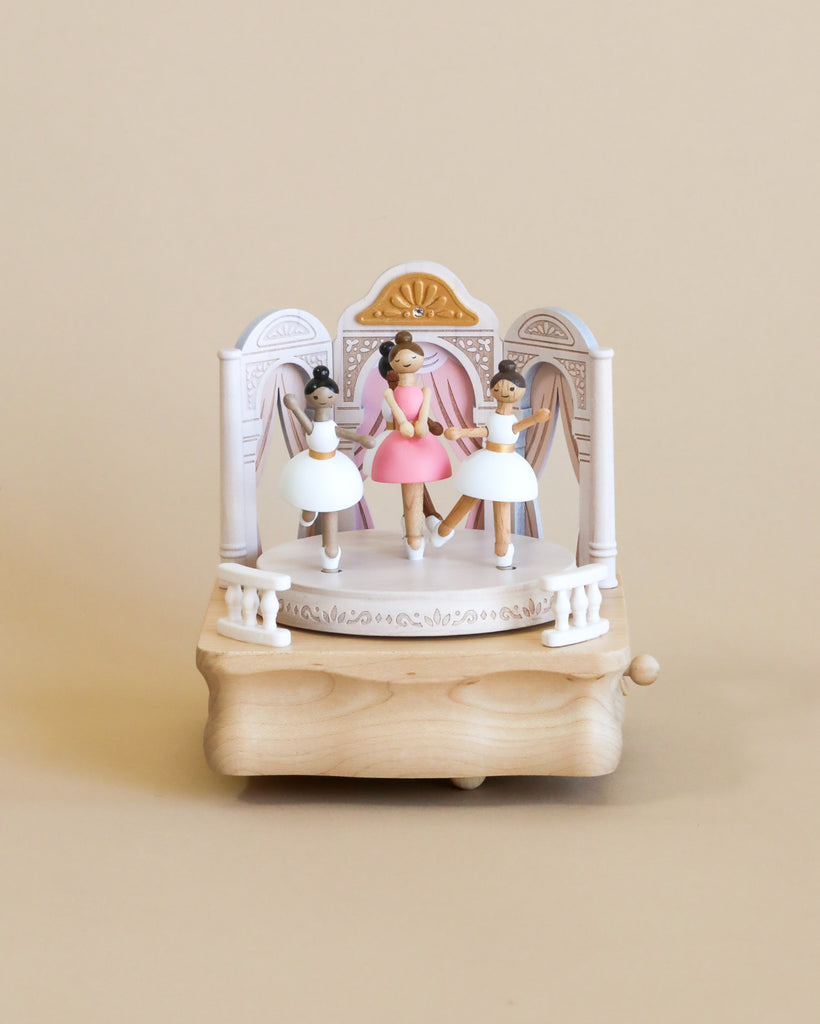 The wooden ballerina music box features a theatrical stage setting, adorned with delicate details. At the center stands a ballerina dressed in a pink dress, and surrounding her are three more ballerinas, each in a graceful stance, wearing white dresses. The ballerinas' features are lovingly painted, showcasing a range of skin tones, adding diversity and depth to the scene.