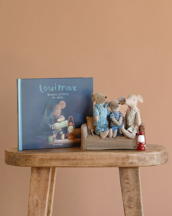 The Maileg LouiMax Storytime Family Set, featuring a children’s book and three mice in pajamas on a couch with a red lantern, is displayed on a wooden table.