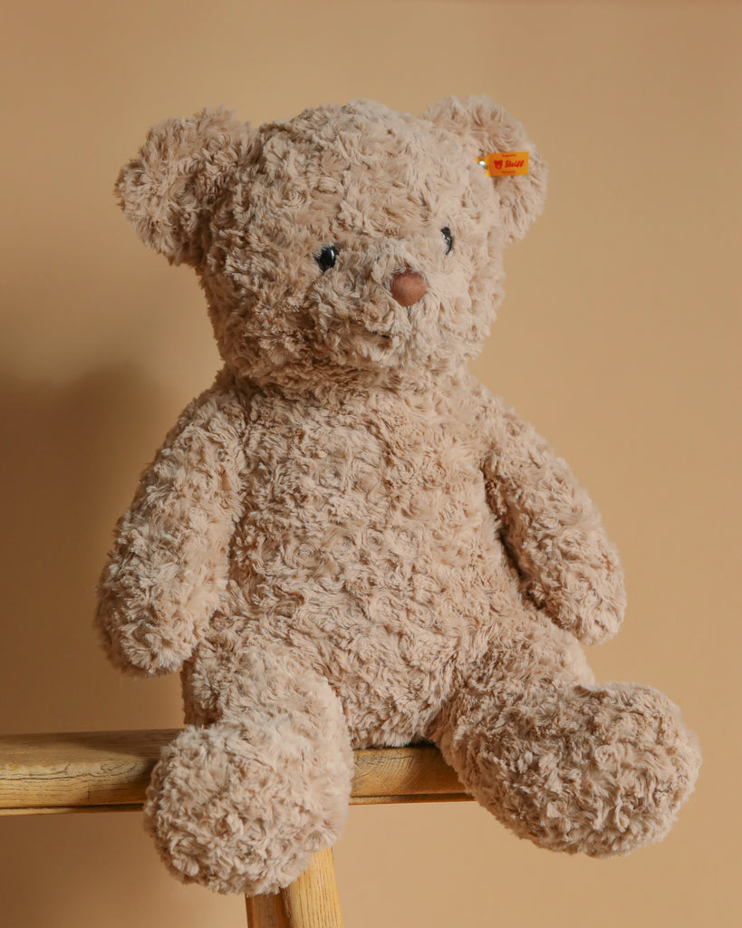 A fluffy, light brown Steiff Honey Teddy Bear, 19 inches tall, is perched on a wooden surface against a beige background. This plush companion has soft fur, small black eyes, a stitched nose, and an orange tag in its ear.