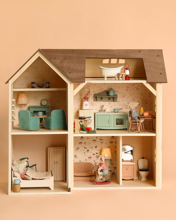 The Maileg Fully Furnished Farmhouse Set + Surprise Gift features pastel-colored vintage-style rooms—kitchen, living room, bedroom, and bathroom—with charming miniature furniture and dolls, all set against a beige backdrop.