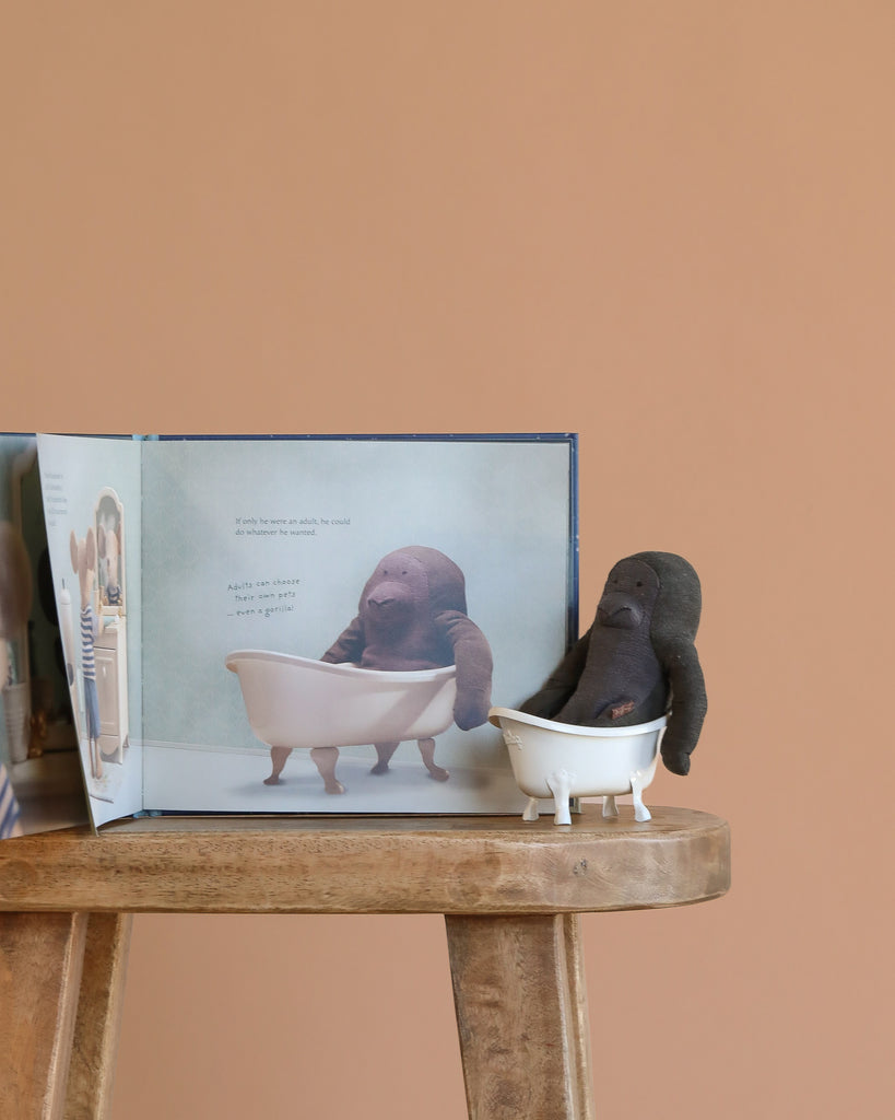 The Maileg Gorilla Bath Time Set is displayed on a wooden stool next to an open book with an illustration of the scene, all set against a plain peach background.