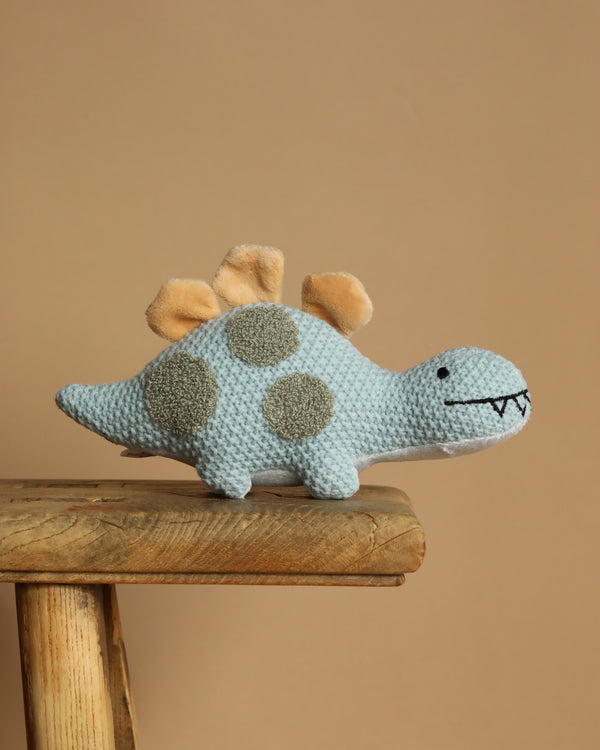 The Baby Stegosaurus Dinosaur Stuffed Animal is an ideal newborn gift, featuring a soft knit with light blue and gray sensory spots and beige fabric spikes. It smiles with embroidered black eyes and mouth while standing on a wooden bench against a plain beige background.