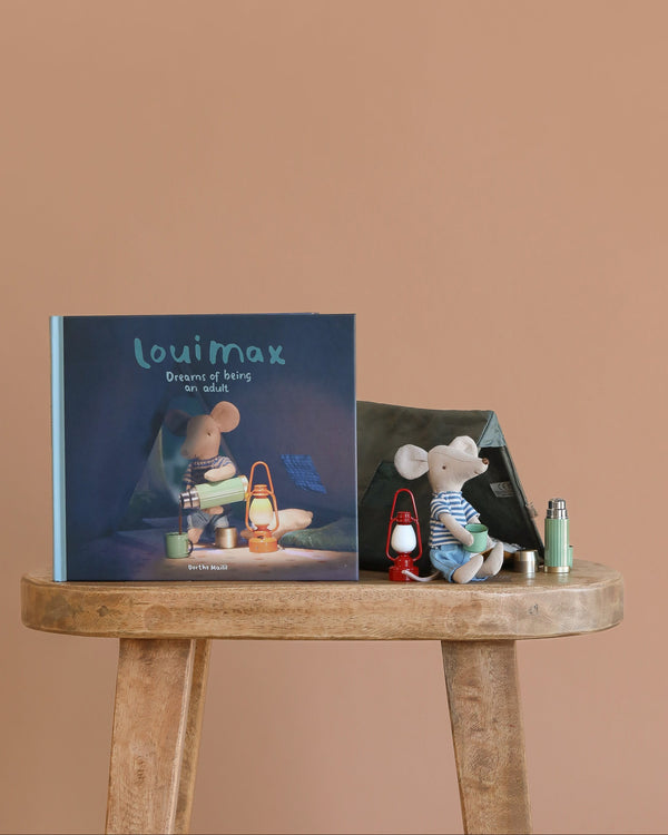 The Maileg LouiMax Little Explorer with Book Set stands on a wooden stool beside a Maileg mouse plush, small tent, lantern, and miniature thermos against a plain beige background.