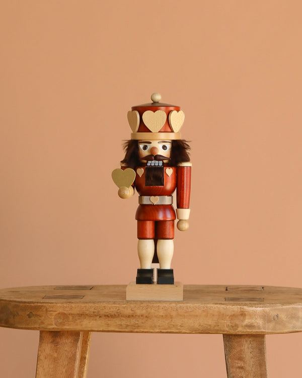 The Christian Ulbricht Nutcracker - Heart King (Natural), made in Germany, stands on a stool against a beige background, wearing a crown and red and gold uniform while holding a gold heart.