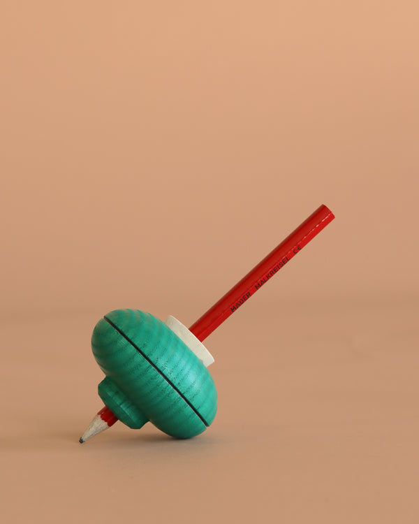 The Mader Malkreisel Drawing Top - Green, a pencil featuring a classic green spinning top at its center, stands upright on a beige surface with a plain beige background.