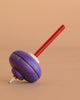 The Mader Malkreisel Drawing Top - Purple, a classic wooden spinning top with a purple body and red handle, rests on its side against a plain beige background.