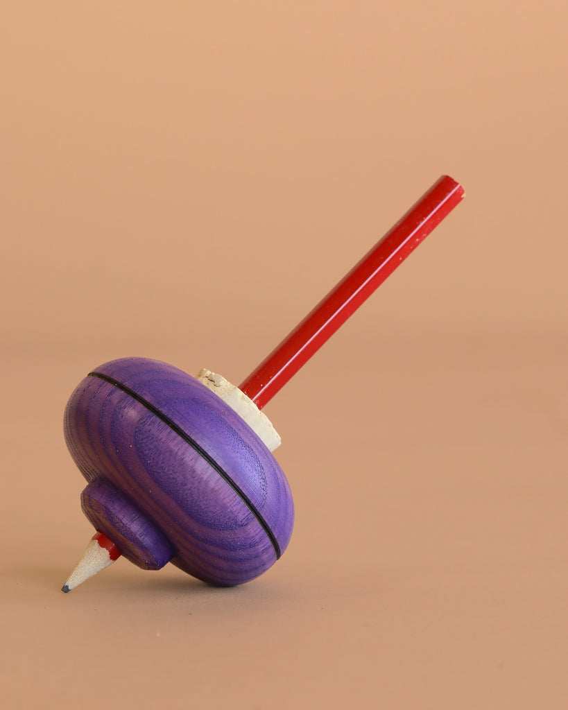 The Mader Malkreisel Drawing Top - Purple, a classic wooden spinning top with a purple body and red handle, rests on its side against a plain beige background.
