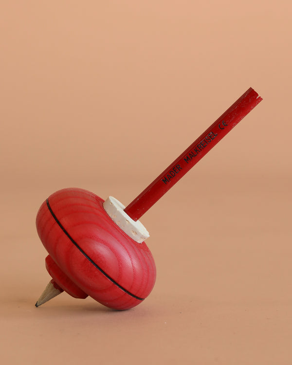 The Mader Malkreisel Drawing Top - Red is a wooden spinning top with a pointed metal tip and stick handle, shown at an angle on a light brown surface against a matching background.