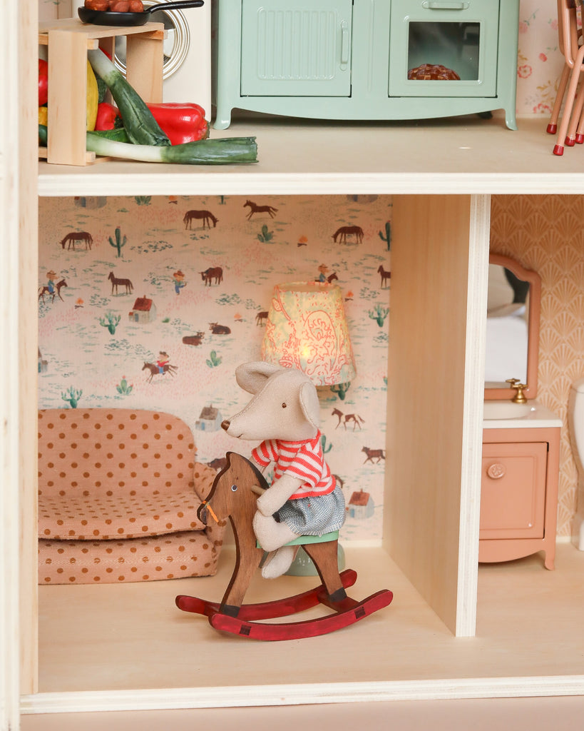 A small mouse doll rides a wooden rocking horse in the Maileg Fully Furnished Farmhouse Set + Surprise Gift, surrounded by miniature accessories including a polka-dot sofa, lamp, and playful horse-and-cacti wallpaper. Ships in 1 week.