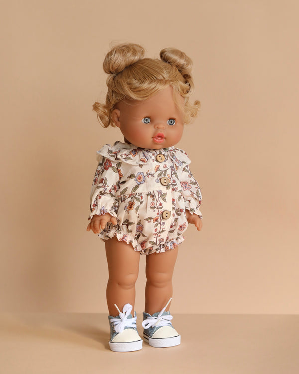 The Minikane Standing Doll (14") - Louisa with Clothing & Shoes features light brown skin, curly blonde hair in two buns, blue eyes, a floral ruffled romper, and white sneakers. This anatomically correct doll is made in Spain and stands against a beige background.