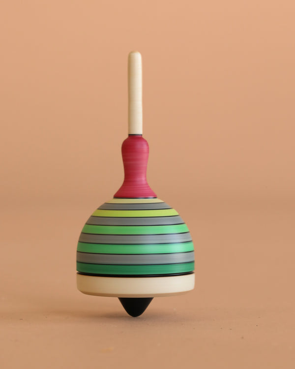 The Mader Fridolin Spinning Top in grey and green stands upright on a beige surface, set against a plain peach background.