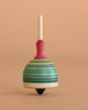 The Mader Fridolin Spinning Top in grey and green stands upright on a beige surface, set against a plain peach background.