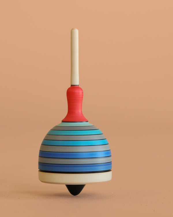 The Mader Fridolin Spinning Top in grey and blue stands upright on a light peach background, showcasing the charm of this classic wooden toy.