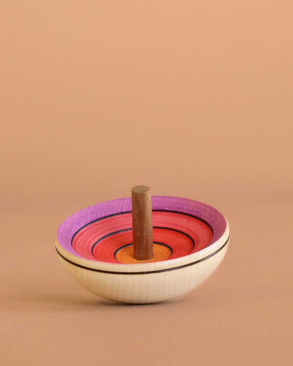 The Mader UFO Spinning Top - Purple Rainbow features vibrant concentric circles in purple, red, and orange with a sturdy wooden handle, standing upright on a light brown surface against a beige background.