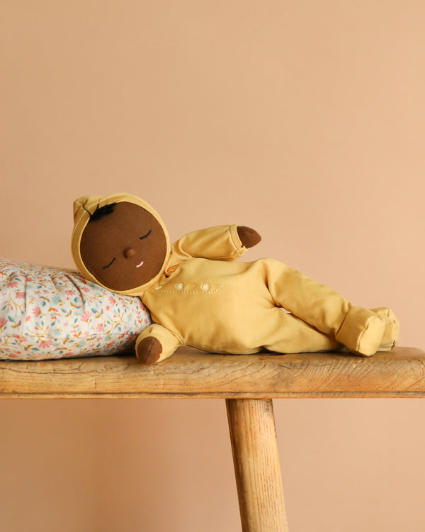A soft brown-skinned Olli Ella Dozy Dinkum - Mini doll in a yellow hood lies on its side, eyes closed, resting on a floral pillow atop a wooden bench against a beige background—a charming keepsake baby toy.