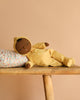 A soft brown-skinned Olli Ella Dozy Dinkum - Mini doll in a yellow hood lies on its side, eyes closed, resting on a floral pillow atop a wooden bench against a beige background—a charming keepsake baby toy.