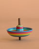 The Mader Sombrero Spinning Top - Striped features vibrant concentric stripes of blue, yellow, red, and green on Maple wood. Crafted in Austria, it stands upright on a light brown surface with a matching beige background.
