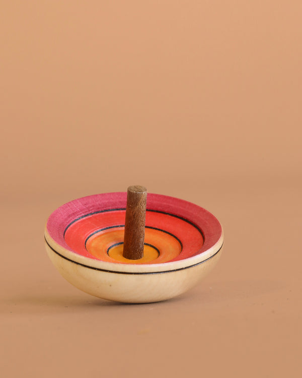 The Mader UFO Flora Spinning Top - Magenta features ombre pink, red, and orange rings with a maple wood handle, displayed on a light brown surface against a matching background.