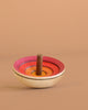 The Mader UFO Flora Spinning Top - Magenta features ombre pink, red, and orange rings with a maple wood handle, displayed on a light brown surface against a matching background.