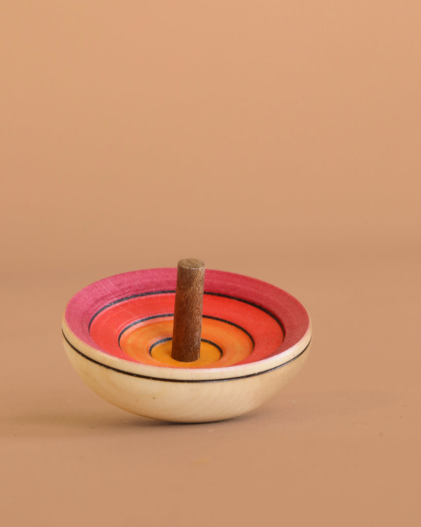 The Mader UFO Flora Spinning Top - Magenta features ombre pink, red, and orange rings with a maple wood handle, displayed on a light brown surface against a matching background.