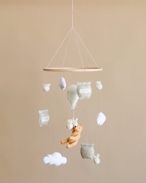 A nursery Handmade Mobile - Honey Bear - Final Sale hanging from the ceiling, featuring a plush teddy bear, white clouds, fluffy sheep, and embroidered cushions with words like "funny" and "happy" on a neutral background.