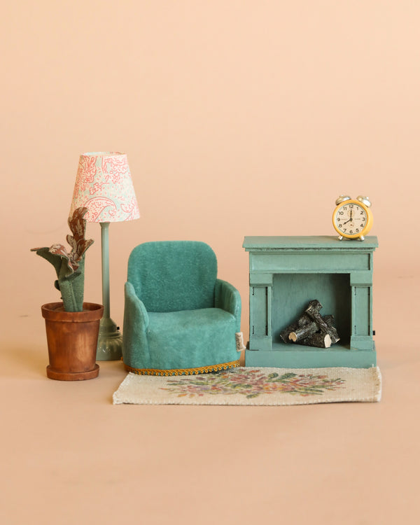 The Maileg Cozy Living Room Set includes a teal armchair, fireplace with logs, floral rug, flower pot, patterned floor lamp, and an alarm clock on the mantel, all styled together on a beige background.