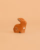 The Bumbu Handmade Wooden Sitting Rabbit is a small, brown, handcrafted figure with white details and a heart on its side, displayed on a solid peach background.