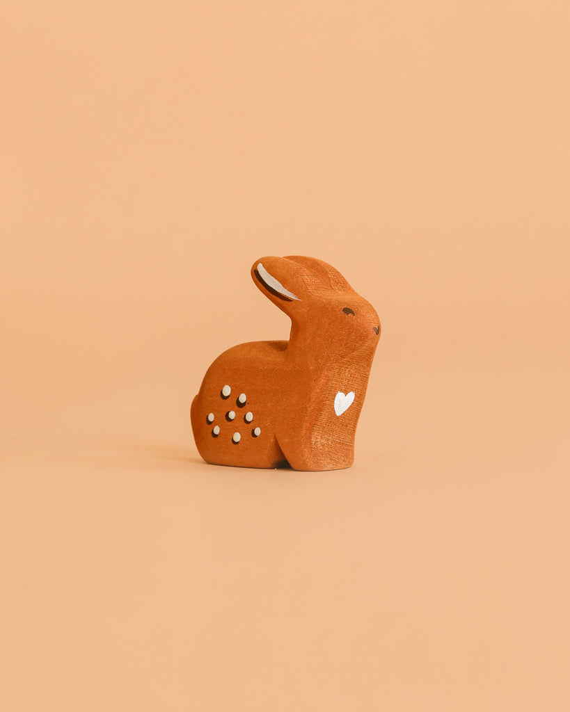 The Bumbu Handmade Wooden Sitting Rabbit is a small, brown, handcrafted figure with white details and a heart on its side, displayed on a solid peach background.