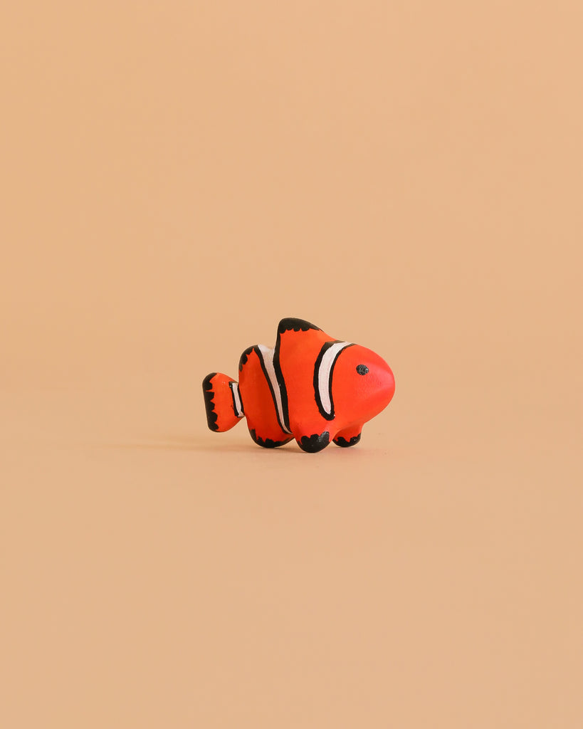 The Bumbu Handmade Wooden Clownfish, featuring orange, white, and black details, is displayed on a peach background. This eco-friendly toy adds minimalistic charm and is ideal for Montessori marine life learning.
