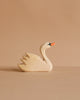 The Ostheimer Handmade Wooden Swan Family Set features small handcrafted wooden swans with orange beaks and simple carved details, displayed on a neutral beige surface against a matching beige background.