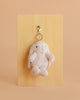 The Snuggle Bunnies - Penelope Keyring - Blossom features a small, plush bunny with floppy ears and a metal clasp, displayed on a wooden board set against a peach-colored background.