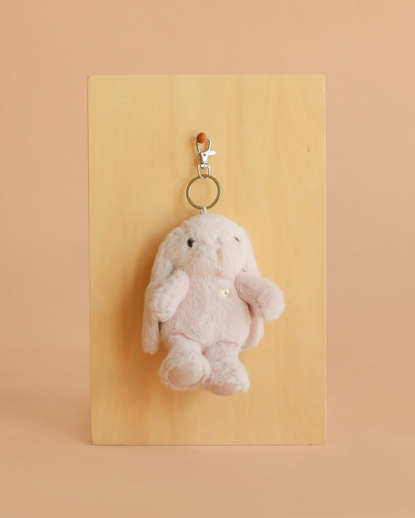 The Snuggle Bunnies - Penelope Keyring - Blossom features a small, plush bunny with floppy ears and a metal clasp, displayed on a wooden board set against a peach-colored background.