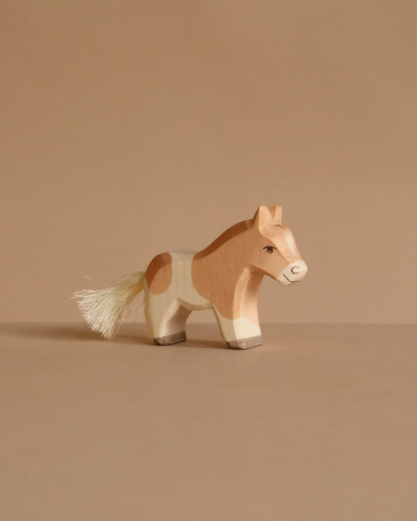 The Ostheimer Shetland Pony Colt, a small wooden toy with light brown and white markings plus a white tail, stands on a beige surface against a beige background, highlighting the charm of handcrafted toys.