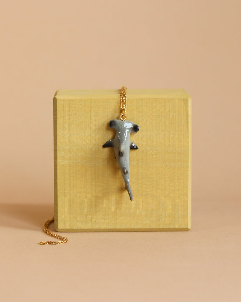 Displayed hanging over the edge of a small wooden cube set against a beige background, the Hammerhead Shark Necklace features a pendant shaped like a gray hammerhead shark on a 24k gold-plated chain.