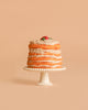 A delightful cake from the Maileg Family Celebration Set, featuring light brown frosting, is elegantly displayed on a white decorative stand against a soft beige background. Topped with a tiny red fruit and green leaf, it captures the whimsical charm characteristic of Maileg accessories.
