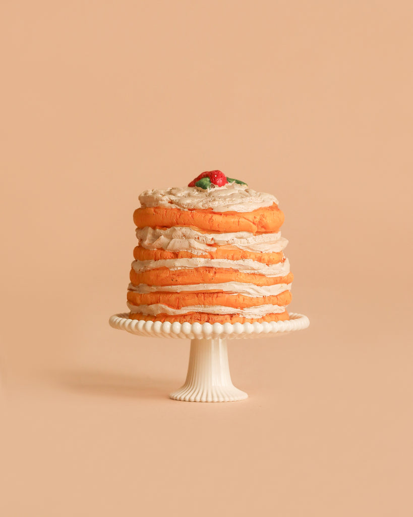 A delightful cake from the Maileg Family Celebration Set, featuring light brown frosting, is elegantly displayed on a white decorative stand against a soft beige background. Topped with a tiny red fruit and green leaf, it captures the whimsical charm characteristic of Maileg accessories.