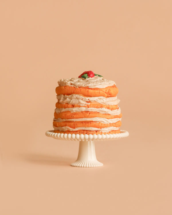 The Maileg Party Cake and Cake Stand, Mouse - Off White features a layered cake with light brown frosting, topped with a small red fruit and green leaves, all set on a white stand. A charming party decoration for your celebration.