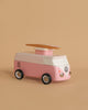 The VW Beach Bus Pink is a pink and white toy van with a wooden surfboard on top, resembling a Volkswagen Type 2, displayed against a plain beige background.